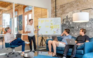 Three men sitting while using laptops and watching man beside whiteboard give employee training