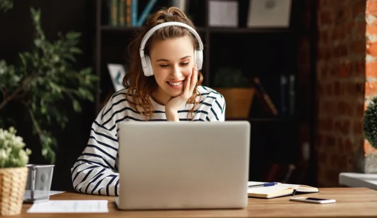 Eine junge Frau mit weißen Kopfhörer sitzt lächelnd vor einem silbernen Laptop und schaut sich eine Online-Infoveranstaltung zu den Studiengängen am Campus der PFH an.