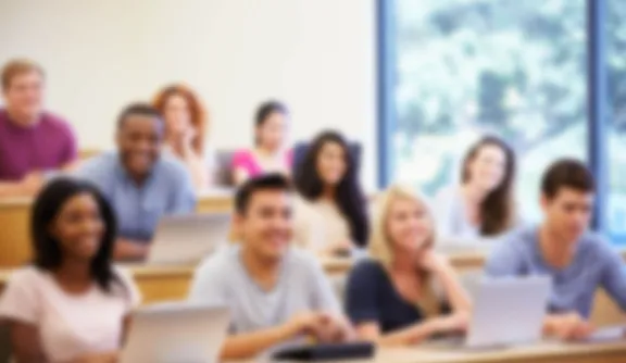 Studierende im MBA Studium an der PFH sitzen in einem hellen Hörsaal und lächeln, ein paar der Studierenden arbeiten an Laptops.